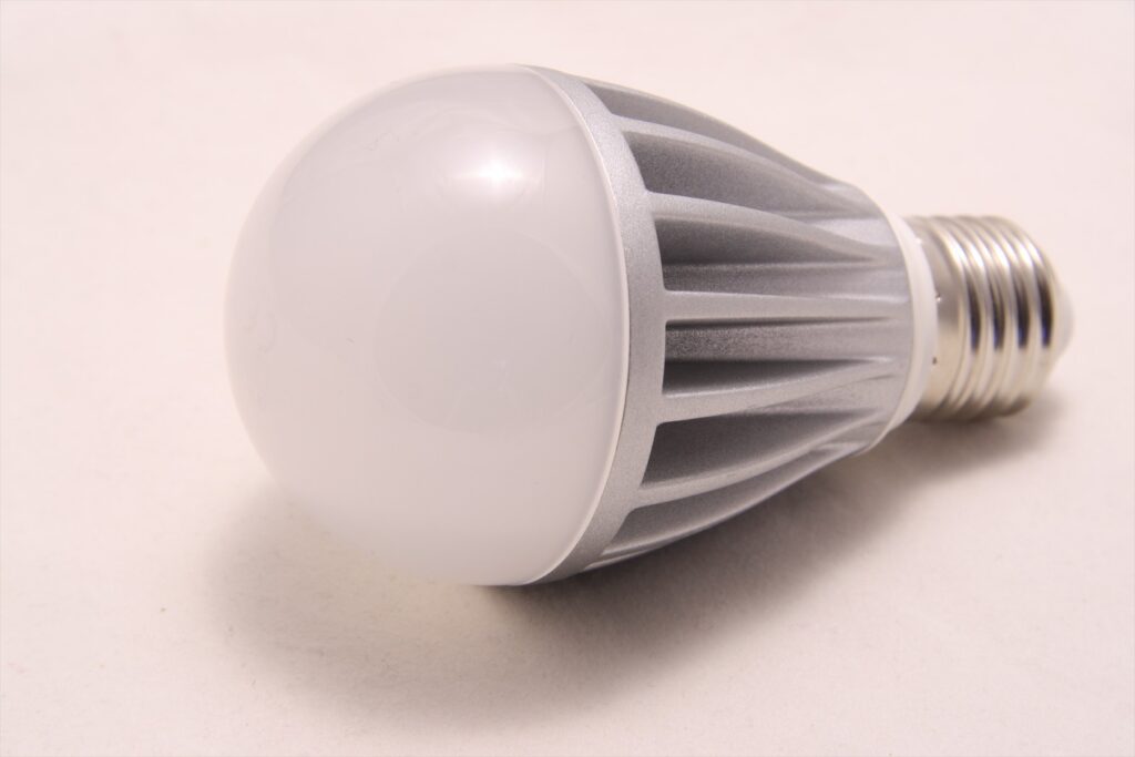 LED žárovka s bílou baňkou, stříbrným žebrovaným tělem a šroubovací paticí na světlém podkladu.