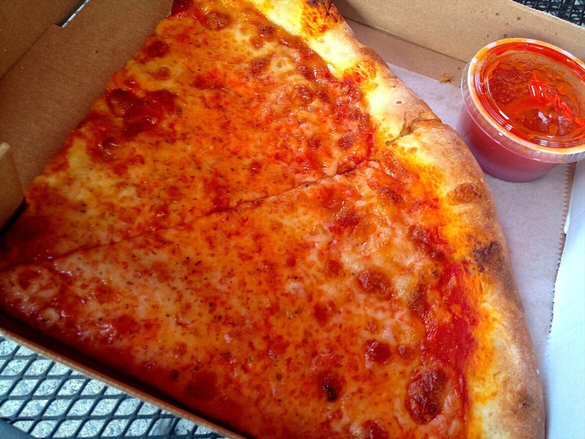 Dva dílky sýrové pizzy v kartonové krabici vedle kelímku s omáčkou.