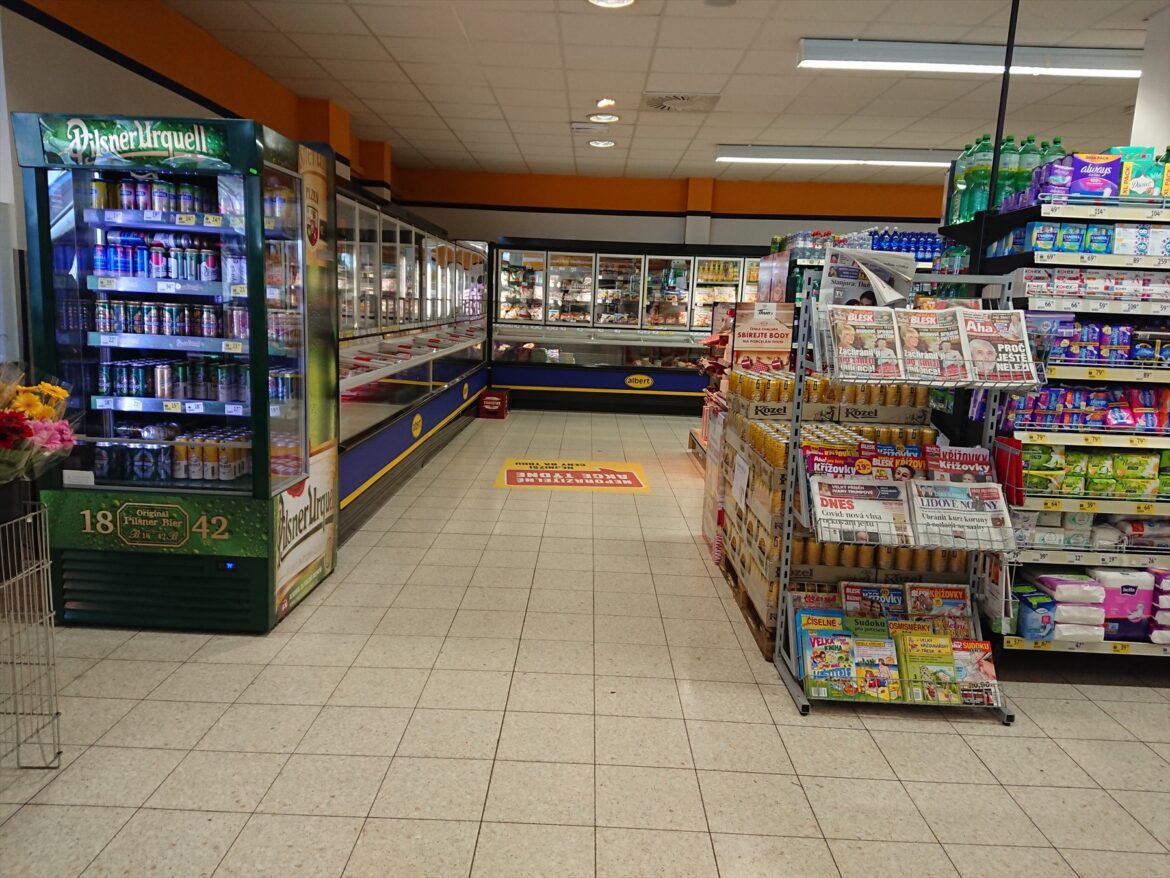 Ulička supermarketu s chladicími boxy, nápoji a stojanem s tiskem.