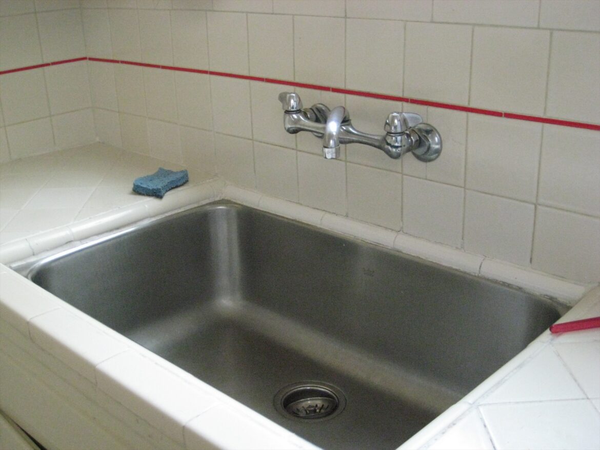 Nerezový kuchyňský dřez pod nástěnnou baterií mezi světlými obklady