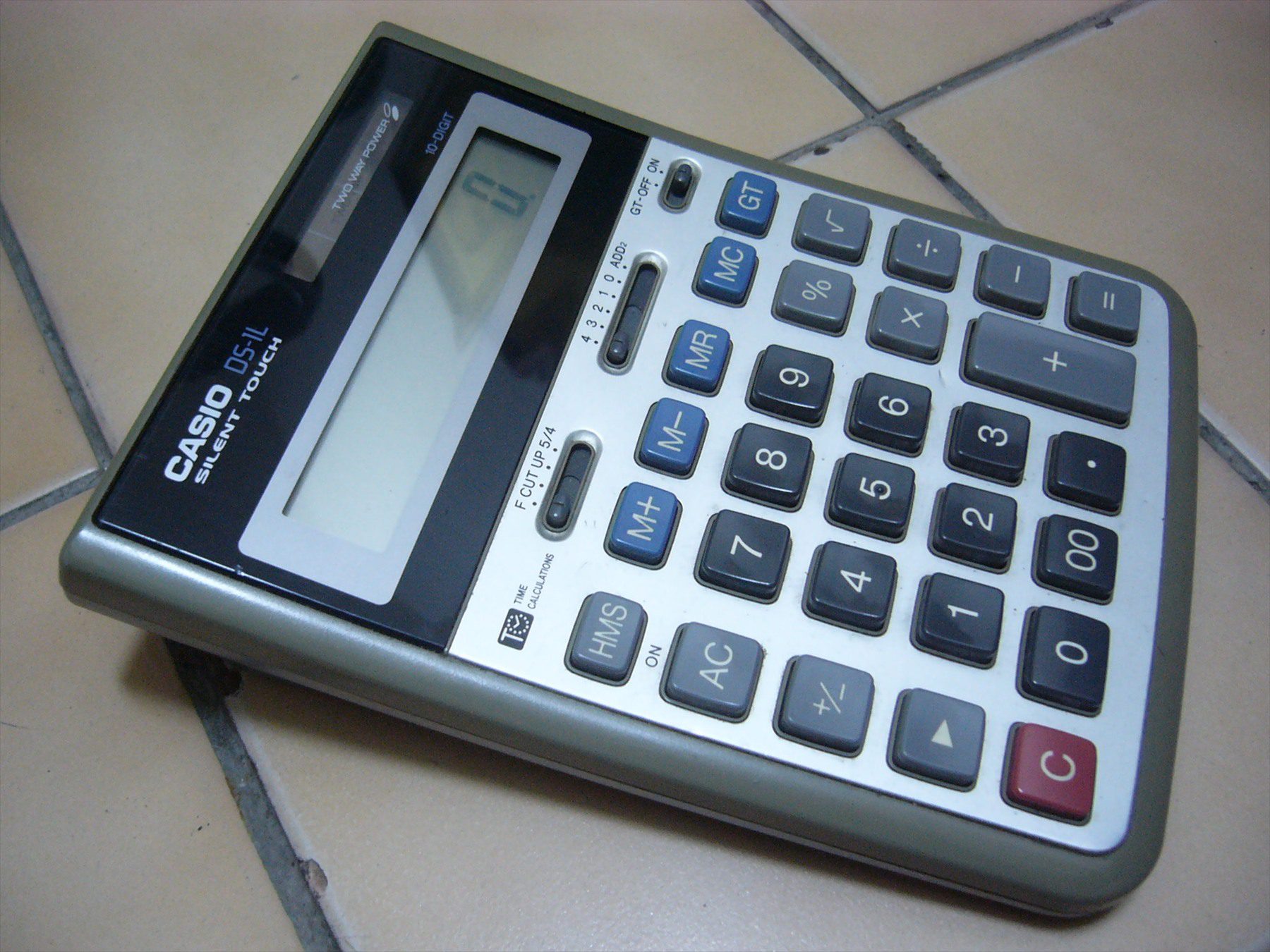 Šedá stolní kalkulačka Casio s velkým displejem položená na dlaždicích.