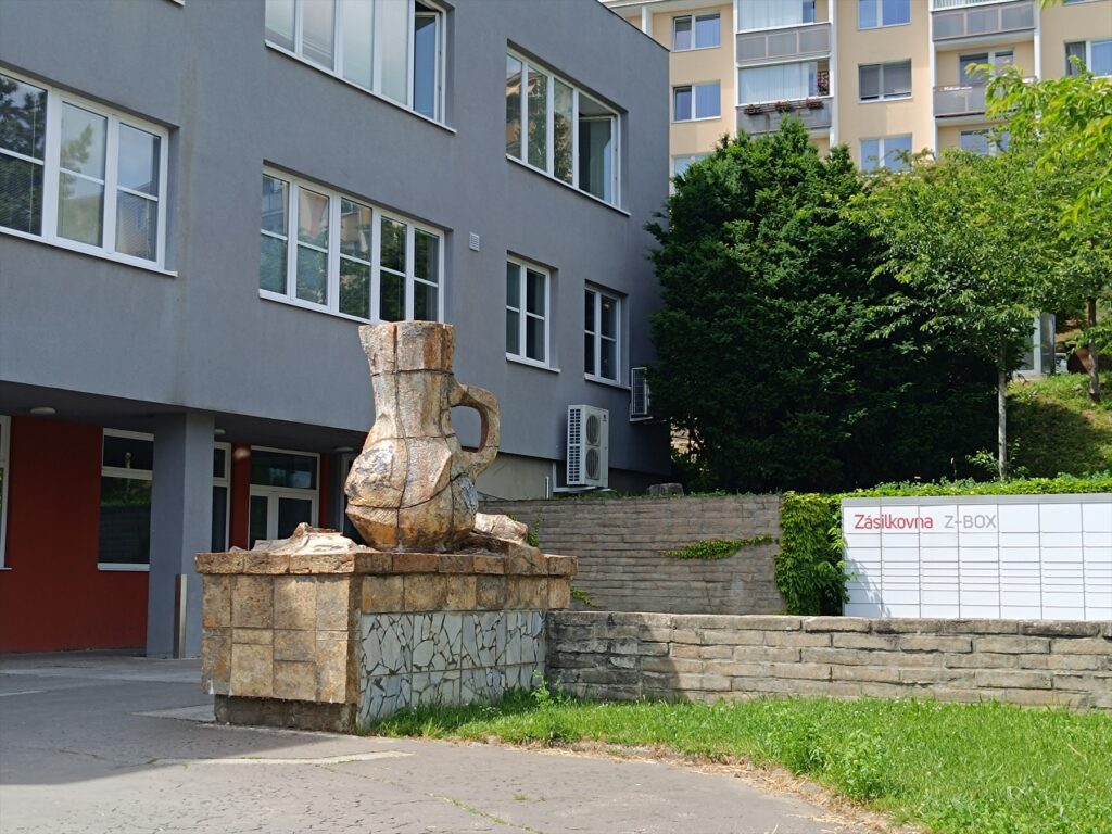 Bílý box Zásilkovny u bytového domu, v popředí kamenná plastika džbánu