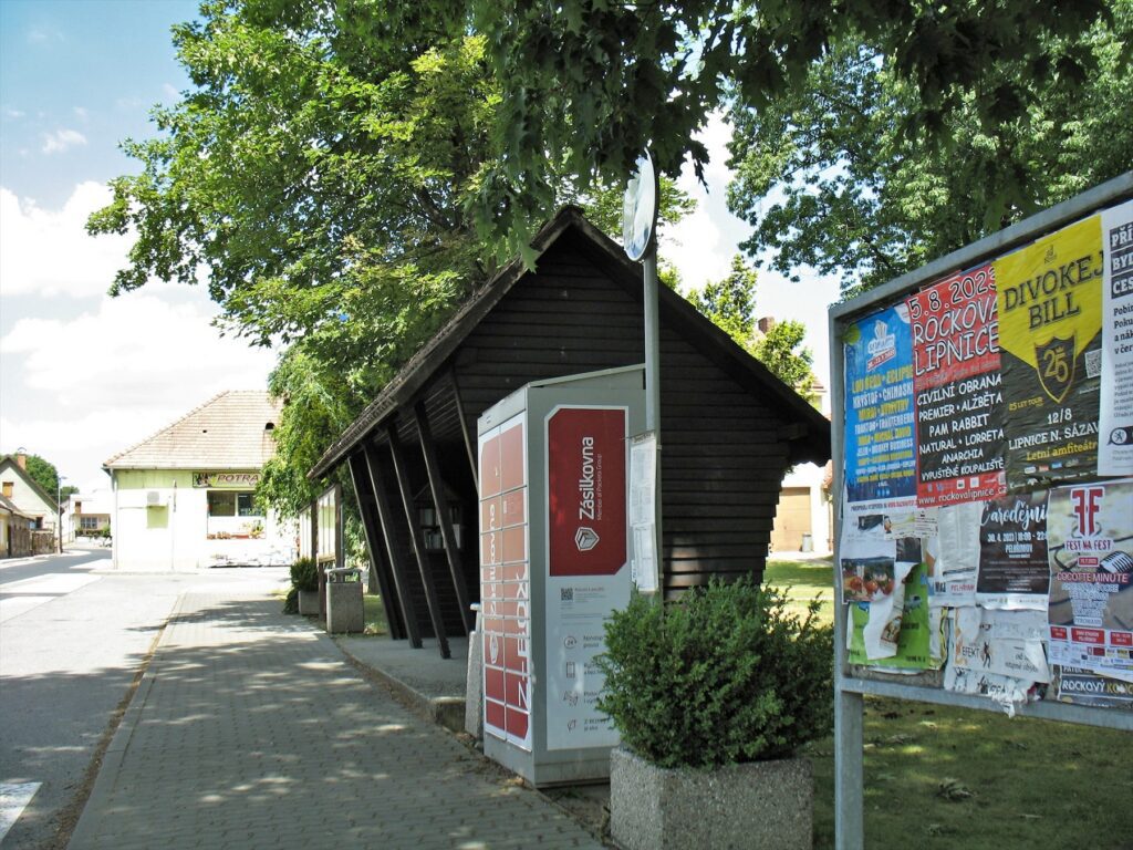 Box Zásilkovny vedle dřevěného přístřešku zastávky a výlepové plochy