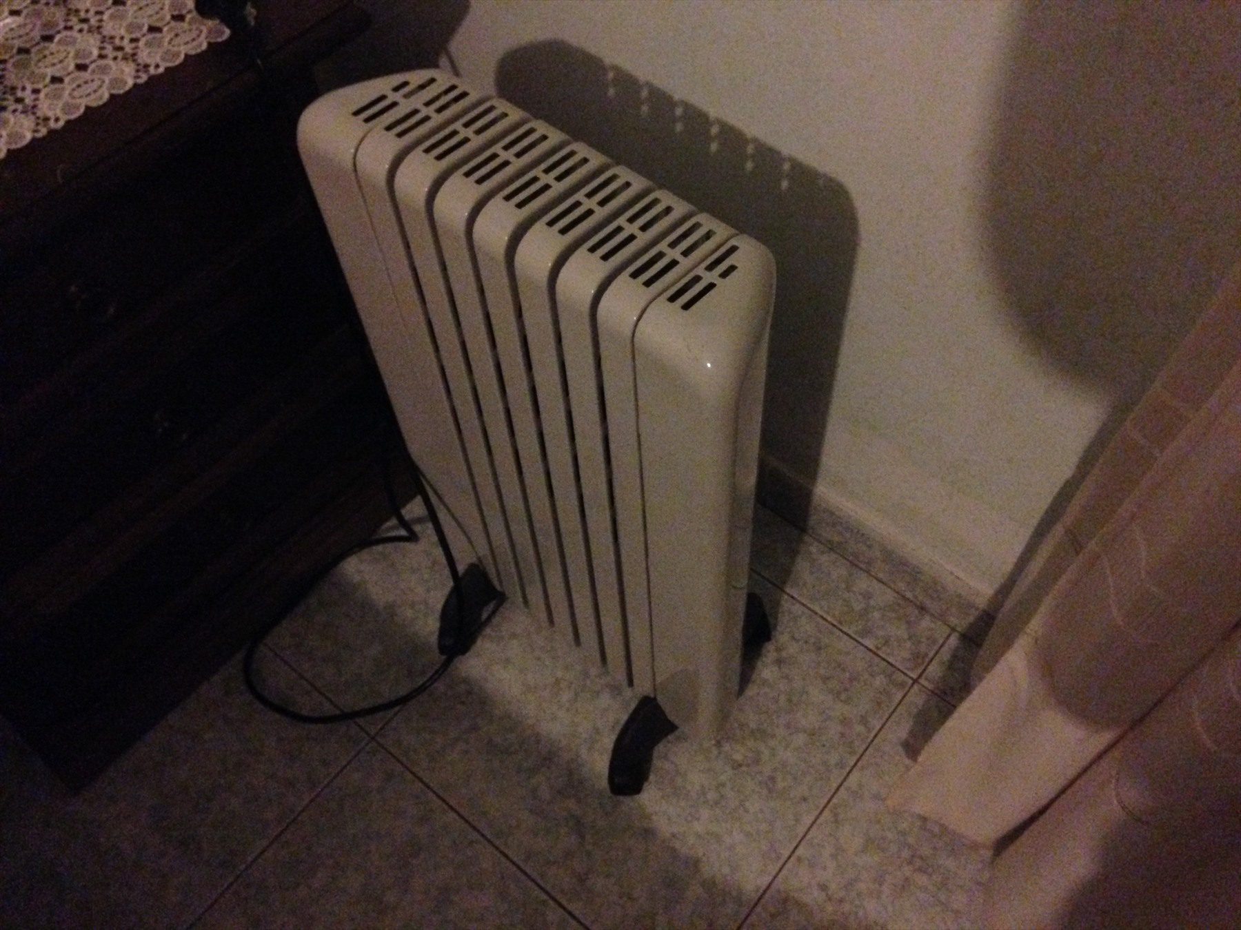 Světlý žebrový olejový radiátor na kolečkách vedle nábytku.