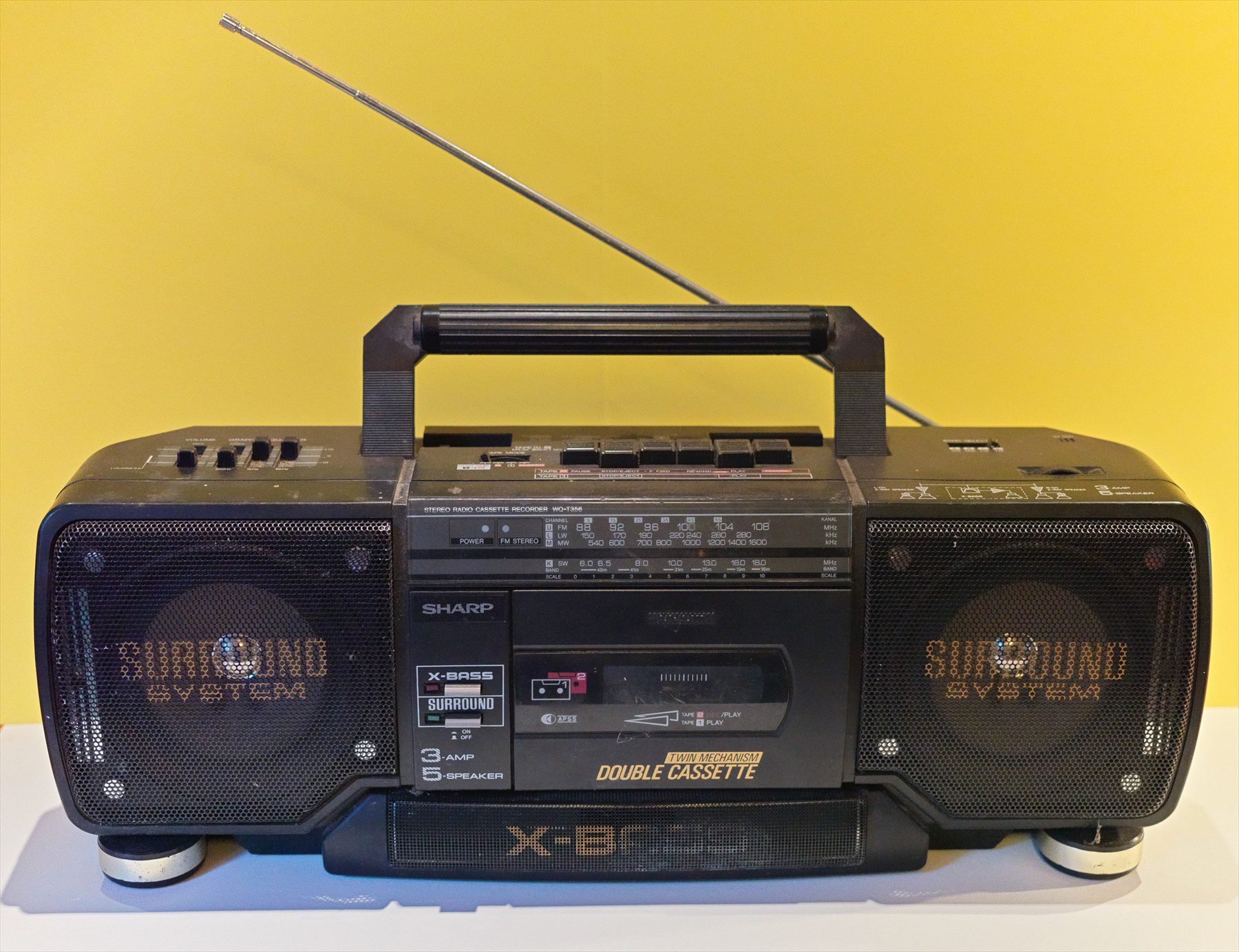 Černý radiomagnetofon Sharp s vytaženou anténou na žlutém pozadí.