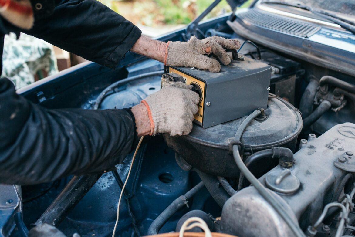 Ruce v rukavicích s měřicím přístrojem u autobaterie v motorovém prostoru.